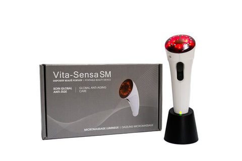 White handle with red end on the Vita-Sensa SM facial stimulator, a handheld device with a sleek, ergonomic design. It features 10 small metal electrodes and 5 red light emitting diodes (LEDs) for microcurrent, light therapy, and vibromassage. The device comes with a gray transformer and base, a user guide, and a portable travel kit.